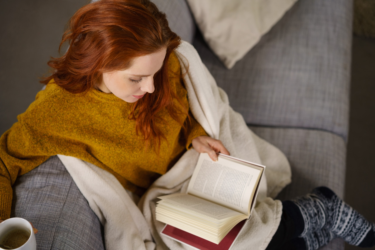 A woman with red hair reading a book on a couch under a cozy blanket