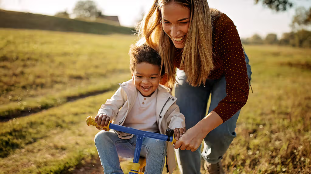 A mother helping her young child ride a bike in a sunny meadow